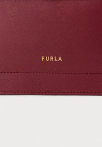 Burgundijas ādas virsma ar tekstūru, kurai ir zelta ieblīvēts "FURLA" logo un kārtīga šūšana gar malām.
