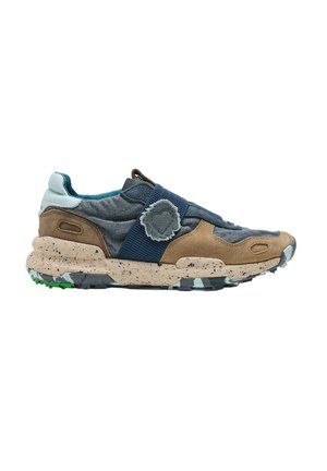 Zapatilla baja con tela azul, paneles de ante marrones, suela beige moteada, suela exterior con estampado de camuflaje y una correa elástica azul con un emblema de tela.