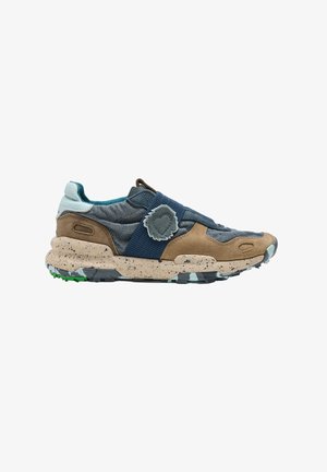 Zapatilla baja con tela azul, paneles de ante marrones, suela beige moteada, suela exterior con estampado de camuflaje y una correa elástica azul con un emblema de tela.