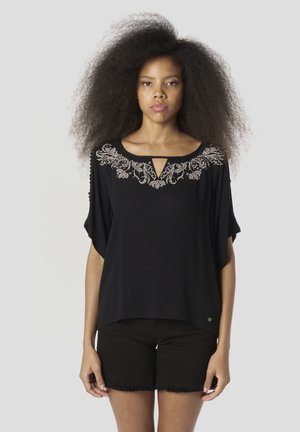 Blusa negra con mangas sueltas, que presenta un intrincado bordado floral blanco alrededor del escote y un pequeño detalle de ojal.