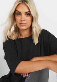 Top nero testurizzato con maniche corte, caratterizzato da righe verticali. La modella ha i capelli biondi di lunghezza alle spalle e un trucco neutro.