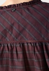 Tissu rayé rouge foncé et gris avec une bordure froncée et un petit bouton rond noir près du bord supérieur.