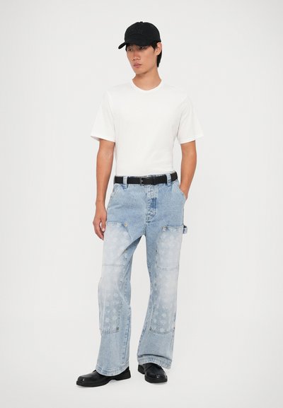 Jeans de mezclilla sueltos de color azul claro con parches en contraste, textura desgastada y bolsillos laterales, combinados con una camiseta blanca básica y una gorra negra.