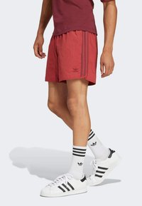 Uomo che indossa pantaloni corti rossi Adidas, calze bianche Adidas con strisce nere e sneakers bianche Adidas, in piedi su uno sfondo semplice.