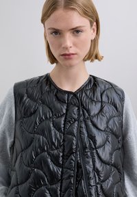 Black quilted puffer vest with a shiny finish, featuring a round neckline, front zipper, and grey sleeves. Textured shell in a fitted design.
