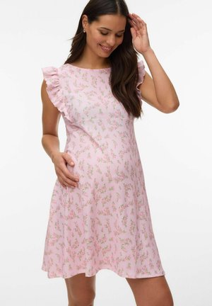 Kobieta w ciąży ubrana w różową kwiatową sukienkę bez rękawów z falbankami na ramionach, delikatnie dotykająca brzucha i uśmiechająca się.