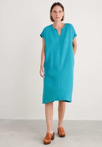 Turquoise, jurk tot de knieën met korte mouwen, subtiele verticale textuur, V-halslijn en zijzakken, gecombineerd met houten clogs met hakken.