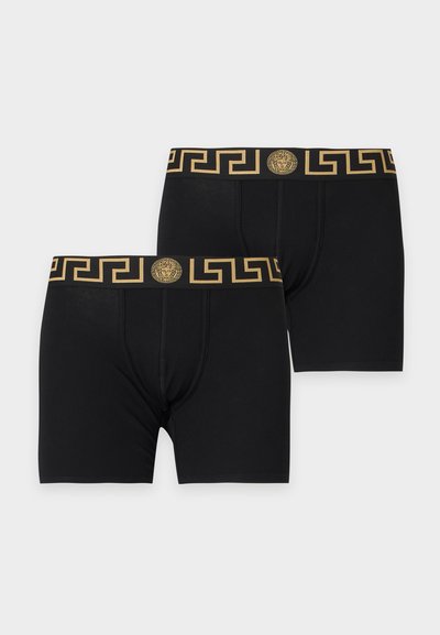 Deux paires de boxers noirs avec motif clé grecque dorée et emblème tête de Méduse sur la ceinture sur un fond clair uni.