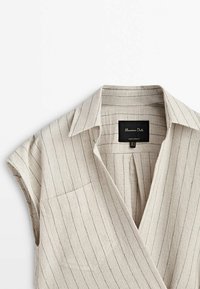 Blusa a righe beige con colletto, maniche corte e taschino sul lato sinistro del petto. Presenta sottili righe nere verticali su un tessuto testurizzato.