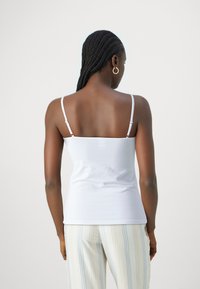 Top branco sem mangas com alças finas e design justo, combinado com calças claras listradas, mostrado de costas. Textura suave.