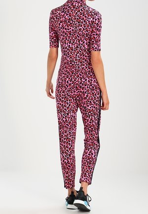 Persoon die een roze en rode, nauwsluitende jumpsuit met luipaardprint draagt, met zwarte zijstrepen en zwarte sneakers met witte zolen, staand met de rug naar de camera.