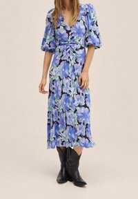 Květinové midi šaty s modrými a zelenými květinami na černém pozadí, s nabíranými rukávy a detailem vázání v pase. Nosené s černými botami.