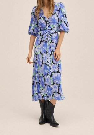 Robe midi florale avec des fleurs bleues et vertes sur un fond noir, munie de manches bouffantes et d'un détail de taille nouée. Portée avec des bottes noires.