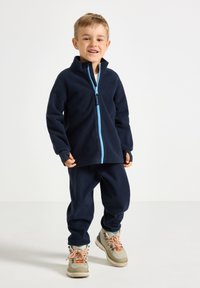 Dunkelblauer Fleece-Jacke mit einem hellblauen Reißverschluss, kombiniert mit passenden Hosen und beigen, wasserdichten Stiefeln mit bunten Schnürsenkeln.