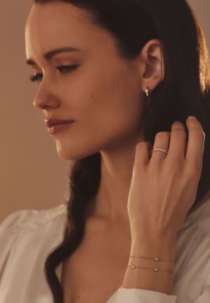 Des bijoux en or portés à l'oreille et au poignet, avec de petits accents ronds. Un bracelet en chaîne délicat avec deux détails semblables à des gemmes.