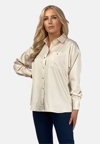 Kremowa bluzka z ostro zakończonym kołnierzykiem, długimi rękawami i kieszonką na piersi. Posiada zapięcie na guziki oraz gładką, błyszczącą fakturę.