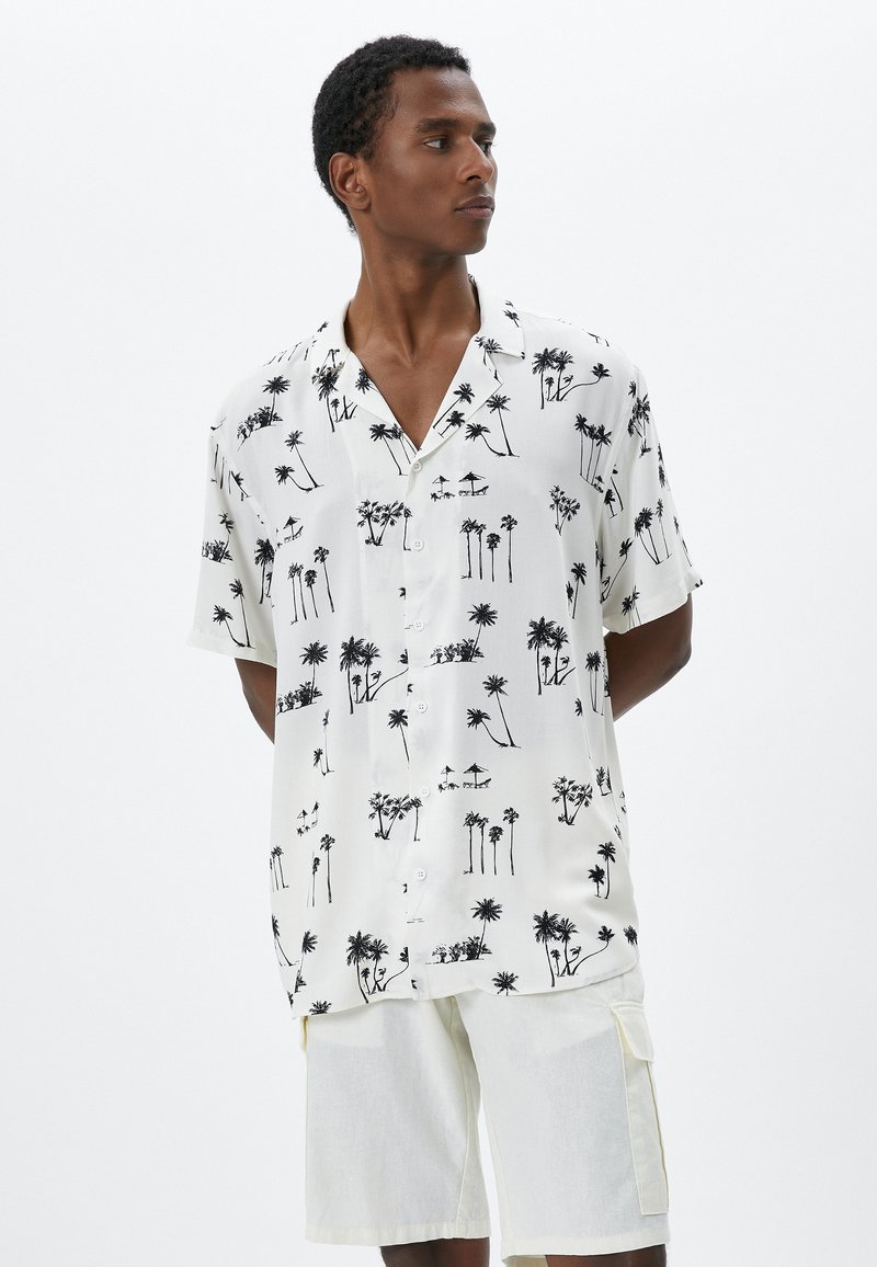 Camicia bianca a maniche corte con stampa di palme nere, dotata di un colletto a scollatura e chiusura con bottoni, abbinata a pantaloni corti beige chiaro.