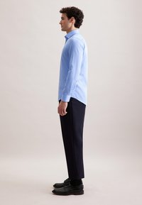 Camicia a maniche lunghe azzurro chiaro con strisce bianche verticali. Abbinata a pantaloni scuri e scarpe nere. La vista laterale evidenzia la vestibilità su misura.