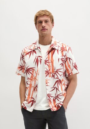 Mężczyzna w białej koszulce pod krótkim, rozpiętym koszulą z nadrukiem czerwonych i pomarańczowych liści bambusa, stoi z rękami w kieszeniach.