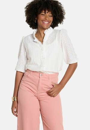 Usmiata žena s kučeravými vlasmi nosí bielu blúzku s otvoreným vzorom a riaseným golierom a ružové nohavice s vysokým pásom, pózujúc s jednou rukou v vrecku.