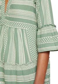 Grön mönstrad klänning med vertikala ränder, geometriska mönster och volangkantade ärmslut. Lätt tyg med strukturerad yta.