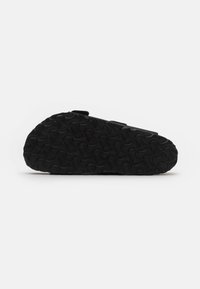 Sandale noire avec une partie supérieure lisse, une sangle réglable et une semelle en caoutchouc texturée présentant un motif en vagues. Offre un design minimaliste.