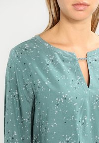 Blusa teal in tessuto morbido, con scollatura a V e piccoli motivi floreali in bianco e nero. Include una cucitura decorativa al collo.