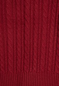 Suéter de punto cableado en rojo profundo, con un patrón texturizado y un dobladillo acanalado. La tela parece suave y cálida, ideal para climas más fríos.