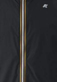 Veste noire à l'avant avec une fermeture éclair centrale argentée bordée de fines bandes rouges et jaunes et un petit logo blanc sur le côté droit supérieur.