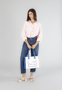 Ljusrosa skjorta med knappar och upprullade ärmar, tillsammans med högmidjade blå denimbyxor och vita klacksandaler med blockklack. Innehåller en vit tote-väska med blå detaljer.
