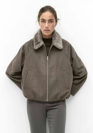 Vrouw draagt een bruine wollen jas met een bontkraag en een rits aan de voorkant, gecombineerd met rechte grijze broek, tegen een effen witte achtergrond.