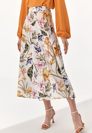 Femme portant une jupe midi blanche à imprimé floral multicolore, associée à un chemisier orange à manches longues et des sandales beiges à talons hauts.