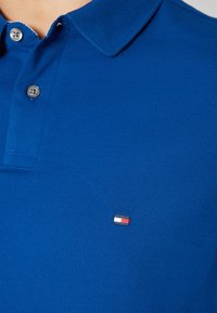 Mėlyna polo marškinėliai, pagaminti iš tekstūruoto medvilnės, su dviem mygtukais ir mažo prekės ženklo logotipu su raudonais, baltais ir mėlynais akcentais.