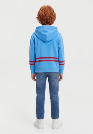 Enfant aux cheveux roux bouclés portant un sweat à capuche bleu avec des rayures rouges, un jean bleu et des baskets blanches, debout de dos sur un fond uni.