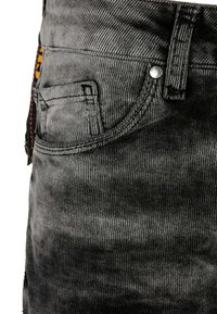 Salopette en denim noir avec une texture côtelée. Comprend une poche avant, un bouton argenté et des accents colorés sur le côté.