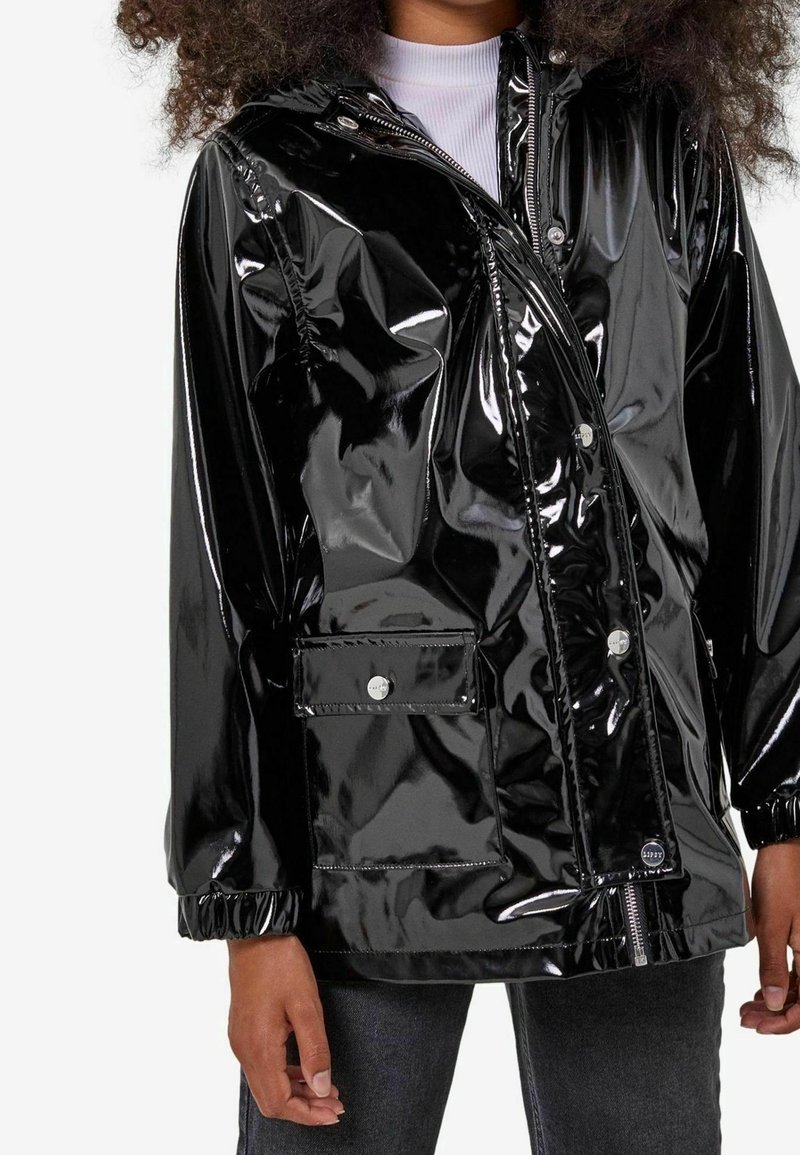 Chaqueta negra, brillante e impermeable con cierre frontal de cremallera, botones a presión plateados, puños elásticos y un bolsillo en el pecho con solapa. Textura suave y brillante.