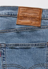 Jeans de denim Levi Strauss con un parche de cuero marrón con texto en relieve. Tejido azul texturizado con detalles de costura amarilla.