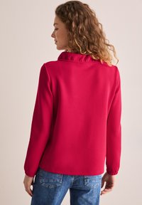Rote Bluse mit rüschigem Kragen, langen Ärmeln und elastischen Bündchen; kombiniert mit blauen Jeans mit Gesäßtaschen. Glatte Textur.