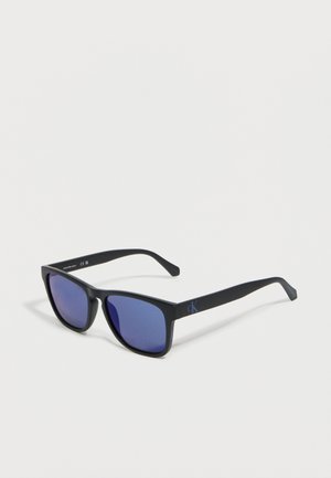 Mustad ristkülikukujulised Calvin Kleini päikeseprillid tumedate siniste läätsedega, mis on asetatud valgele pinnale.