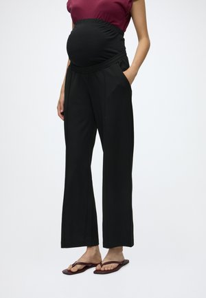 Personne enceinte portant un pantalon de maternité noir, un haut bordeaux sans manches et des sandales marron, debout avec une main dans la poche.