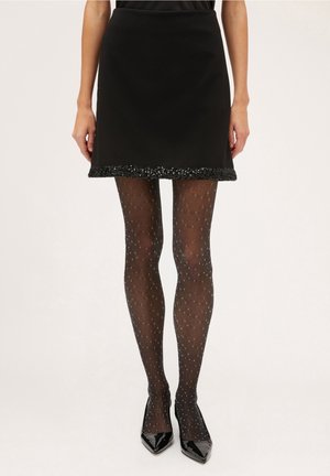Mujer con minifalda negra con dobladillo de lentejuelas, medias negras transparentes con estampado y tacones negros brillantes de punta afilada sobre un fondo blanco.