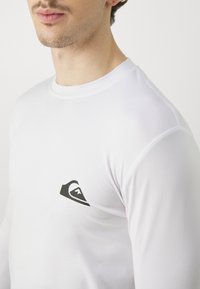 Balta ilgās piedurknes krekls, izgatavots no gludas auduma, ar nelielu melnu logotipu augšējā kreisajā pusē. Apaļš kakla izgriezums, pieguļoša piegriezuma dizains.