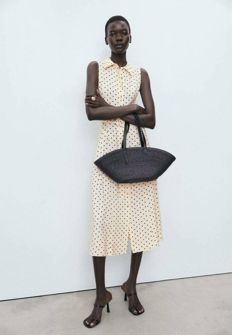 Vestido de lunares amarillo claro con mangas cortas, cierre de botones en la parte delantera y hasta la rodilla. Bolso de mano negro tejido con forma curvada y doble asa.