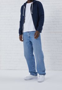 Uomo che indossa una felpa blu con zip, una maglietta bianca, jeans azzurri chiari e sneaker bianche mentre si appoggia a un muro di mattoni bianchi.