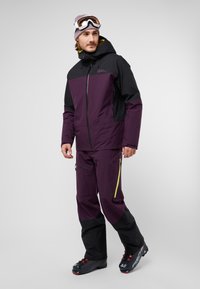 Outfit da sci con giacca nera e viola con zip frontale, abbinata a pantaloni viola coordinati e scarponi da sci neri. Accenti di zip verdi notevoli.