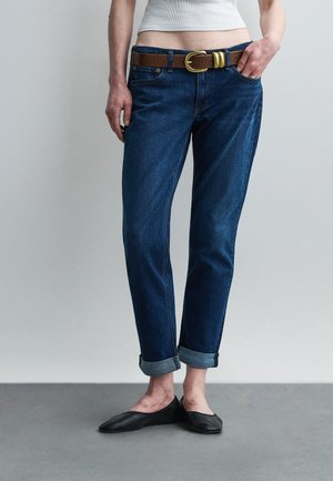 Άτομο που φοράει σκουρόχρωμα μπλε τζιν με γυρισμένο στρίφωμα, καφέ ζώνη και μαύρα επίπεδα παπούτσια, στέκεται μπροστά σε ένα απλό ανοιχτό γκρι φόντο.