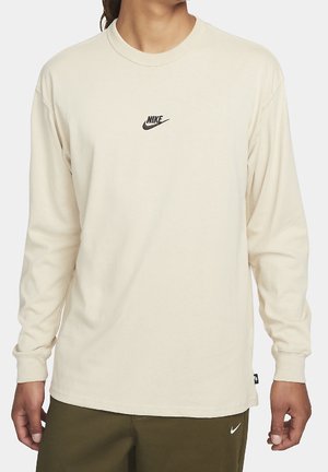 Person trägt ein beiges Nike-Langarmshirt mit kleinem schwarzem Logo auf der Brust, kombiniert mit olivgrünen Hosen mit kleinem weißem Nike-Swoosh.