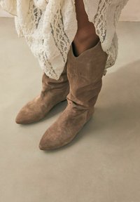 Botas de ante marrón hasta el tobillo con punta afilada, diseño arrugado, superficie texturizada y un sutil detalle de costura.