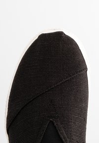 Gros plan sur une chaussure noire en tissu à enfiler avec une semelle blanche en caoutchouc et une couture diagonale sur le dessus.