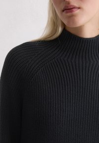 Pull en maille noire avec un col montant, présentant une texture côtelée et des détails de couture aux épaules. Le tissu a l'air épais et chaud.